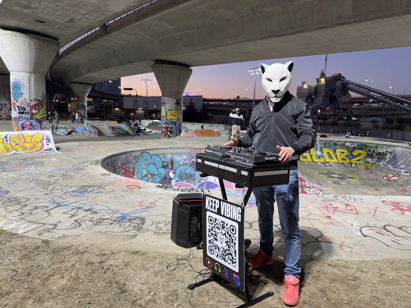 Agu Ocha wearing the geometric mask behind DJ decks at a skate park